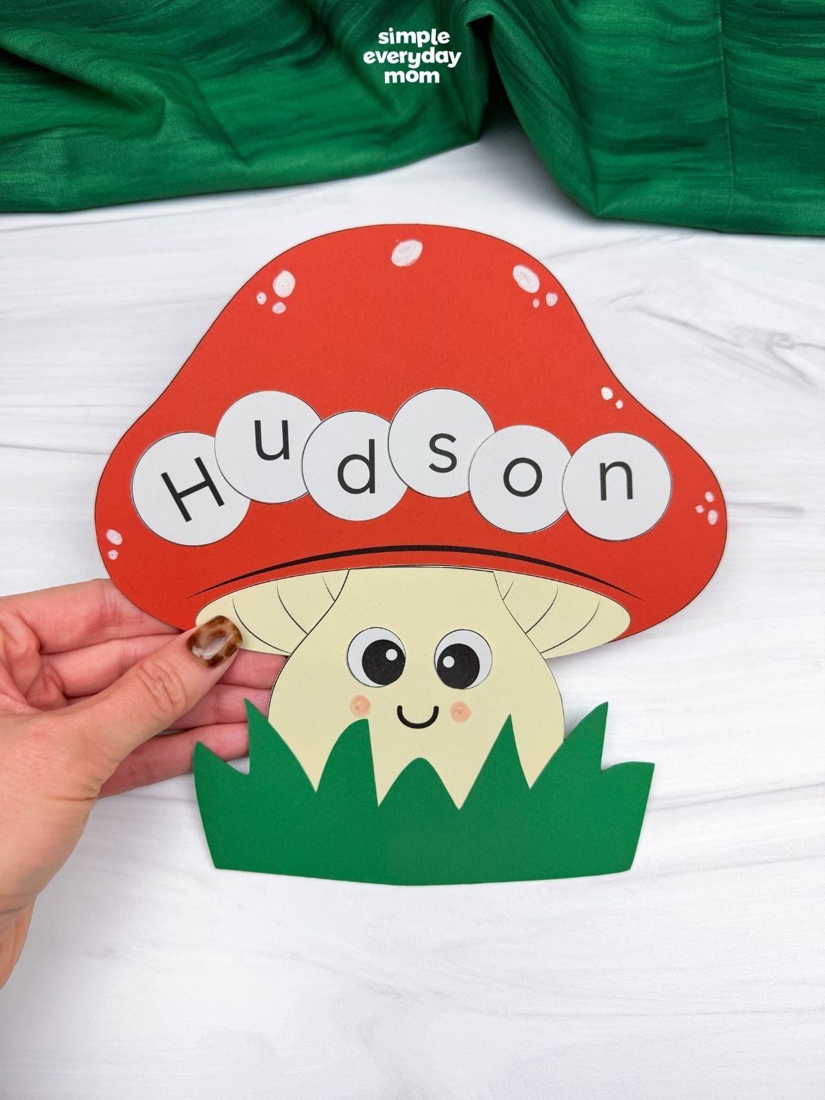 Hands holding a completed mushroom name craft with an orange cap spelling Hudson and green grass.