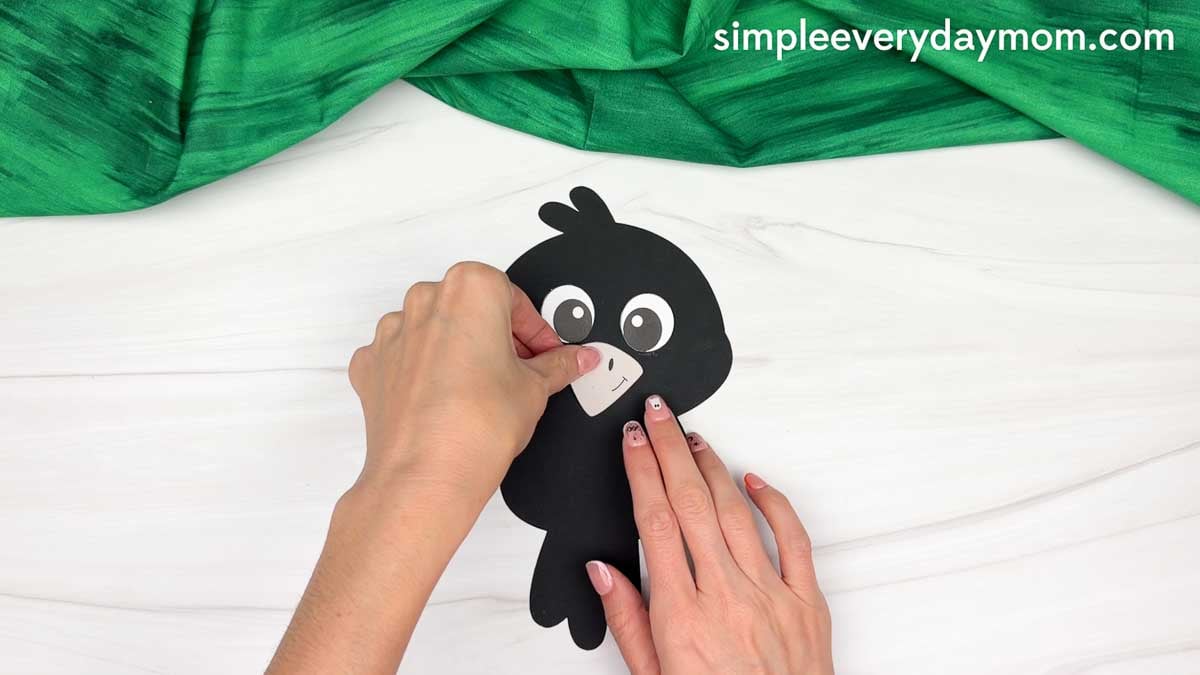 Hands gluing the beak below the eyes on the crow’s face.
