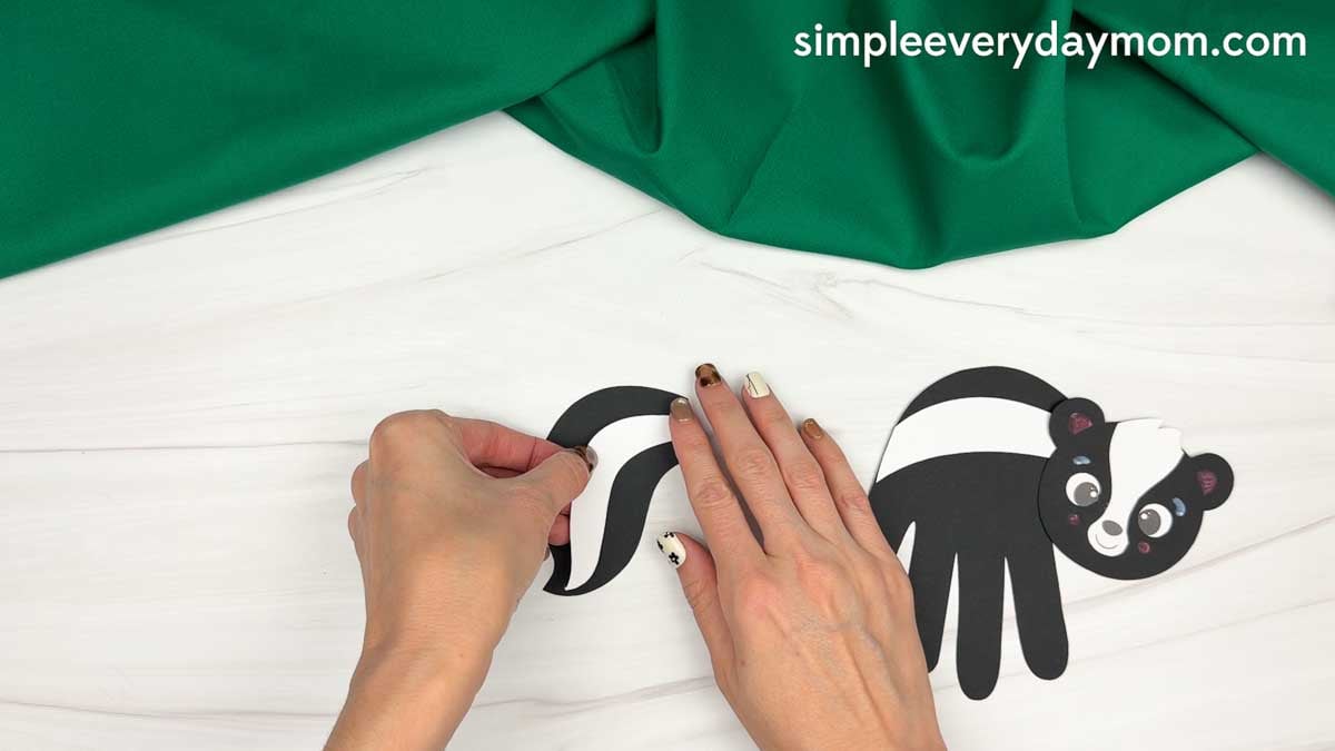 hands assembling the black and white skunk tail beside the handprint skunk craft.