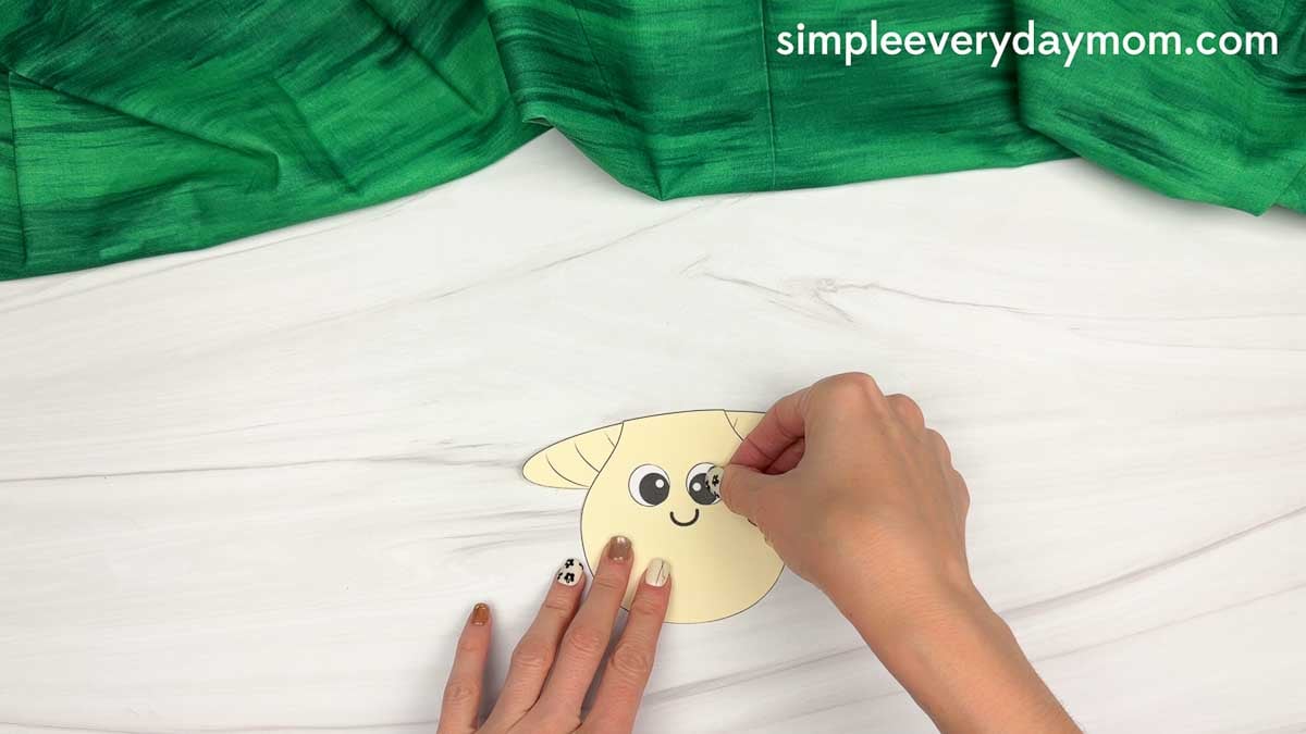 Hands attaching large eyes to the smiling beige mushroom body for the mushroom name craft.