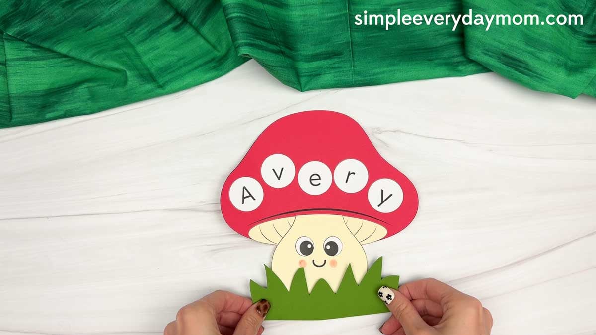Hands adding green grass to the bottom of the mushroom name craft with “Avery” on the red cap.