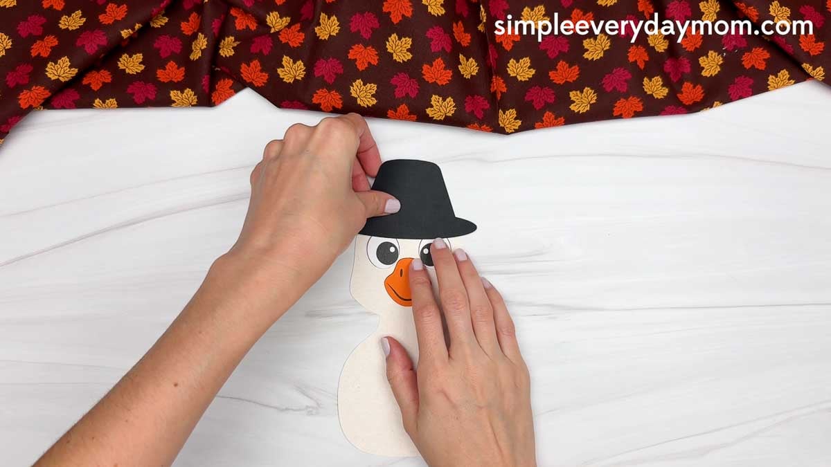 Hands gluing a black pilgrim hat onto the goose's head for a Thankful Goose Craft.