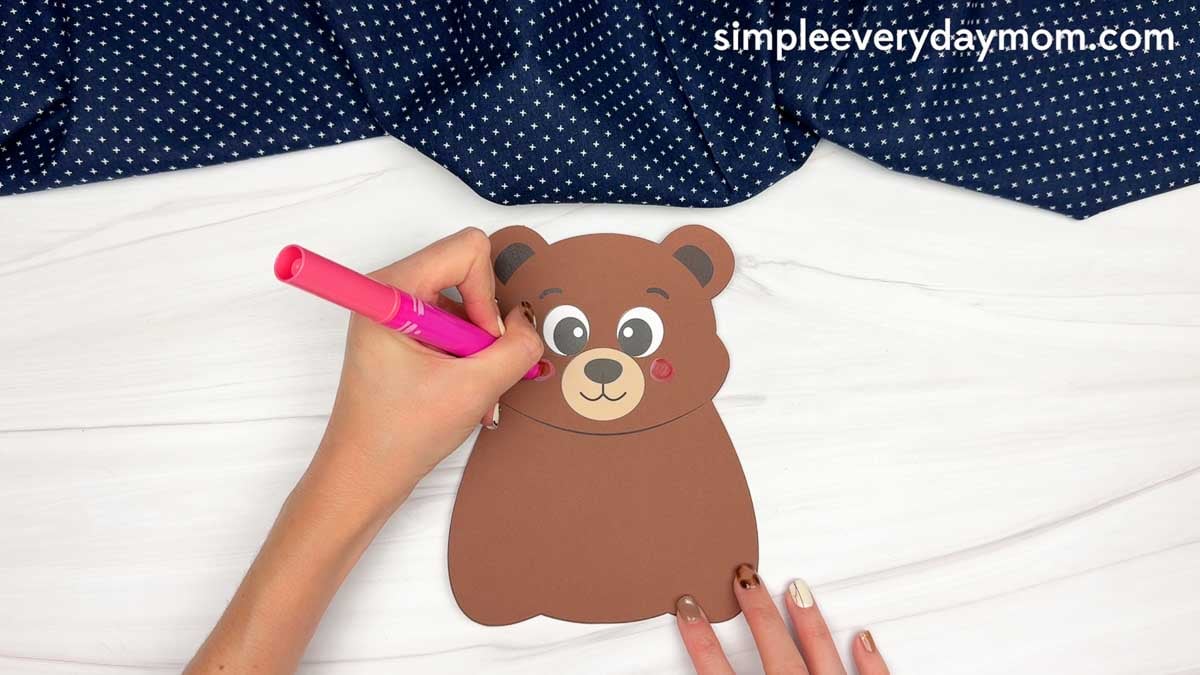 hands coloring pink cheeks on a brown paper bear craft with a marker.
