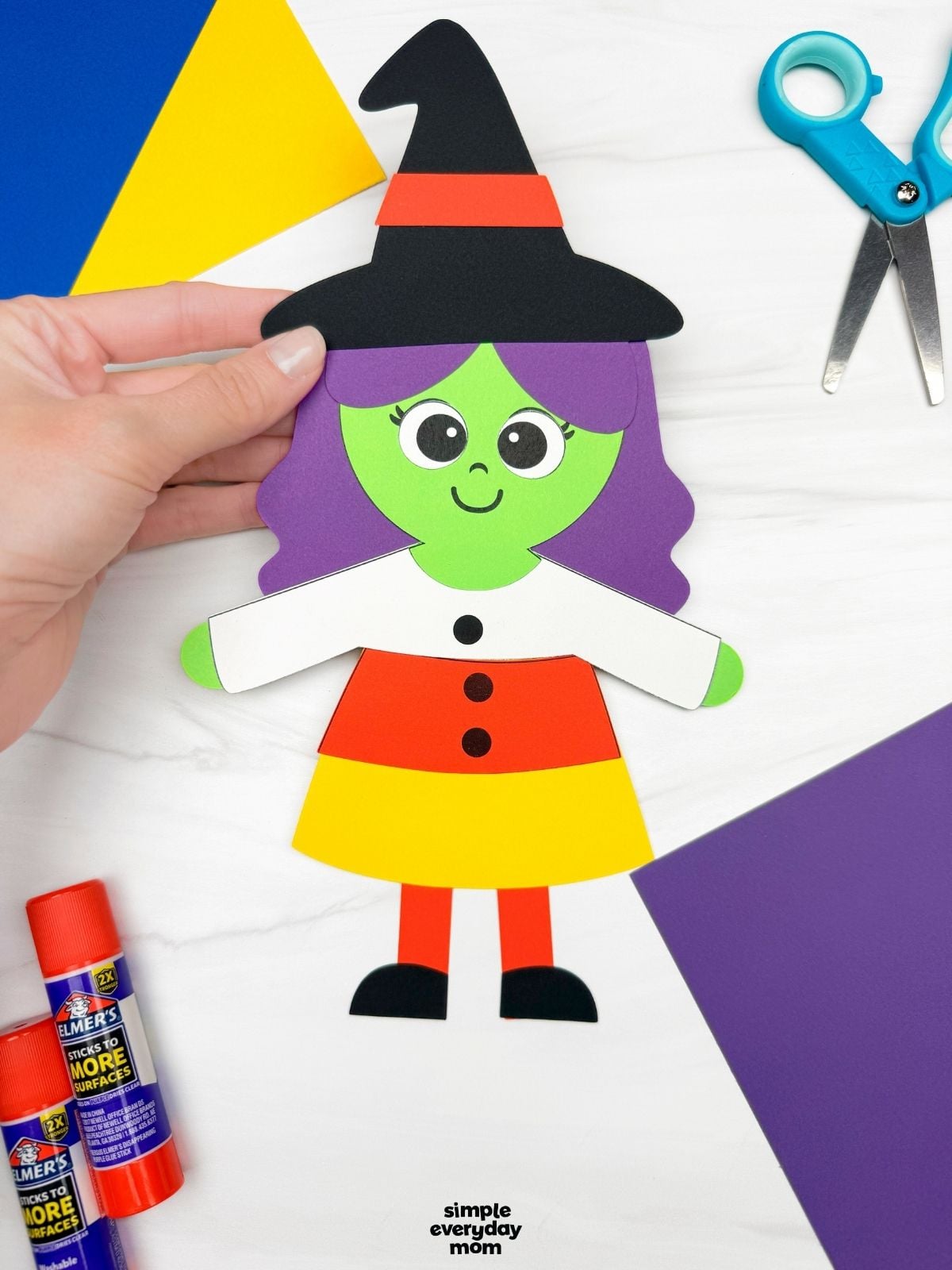 Hands holding a completed Witch Candy Corn Craft with purple hair, a witch hat, and a candy corn dress.