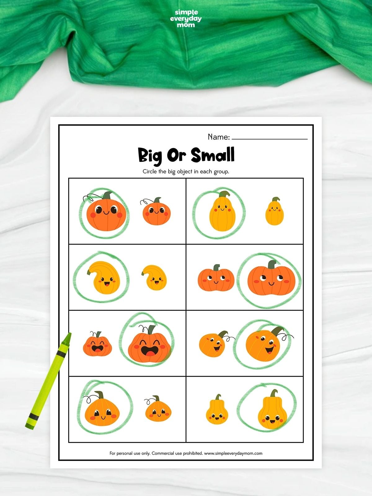 A completed big or small pumpkin worksheet shows pairs of cheerful pumpkins and gourds in different sizes. The larger object in each pair is circled in green for a simple size-comparison activity. A green crayon rests beside the printable on a white surface with green fabric above.