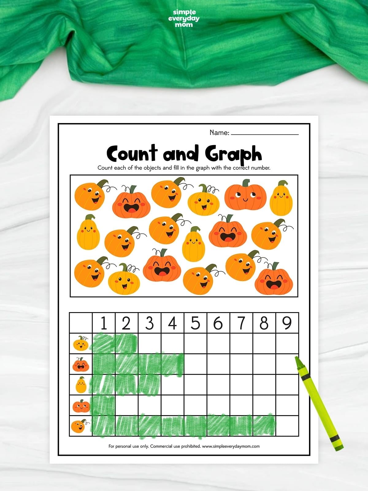 A completed pumpkin count and graph worksheet features several cheerful pumpkin and gourd designs to count. A green bar graph below records how many of each design appear in the picture. A green crayon rests beside the printable on a white surface with green fabric above.