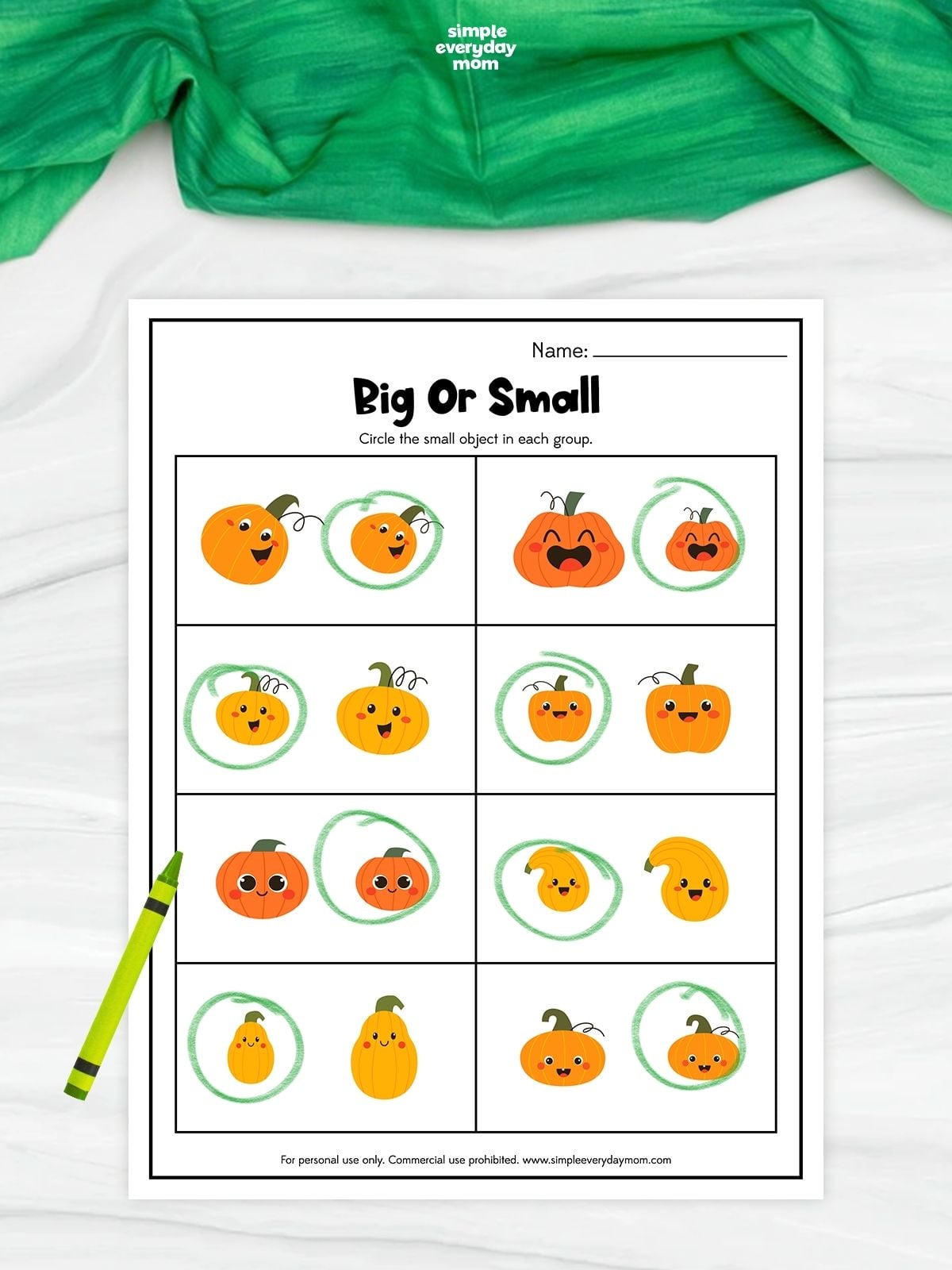 A completed big or small pumpkin worksheet shows pairs of cheerful pumpkins and gourds in different sizes. The smaller object in each pair is circled in green for a simple size-comparison activity. A green crayon rests beside the printable on a white surface with green fabric above.