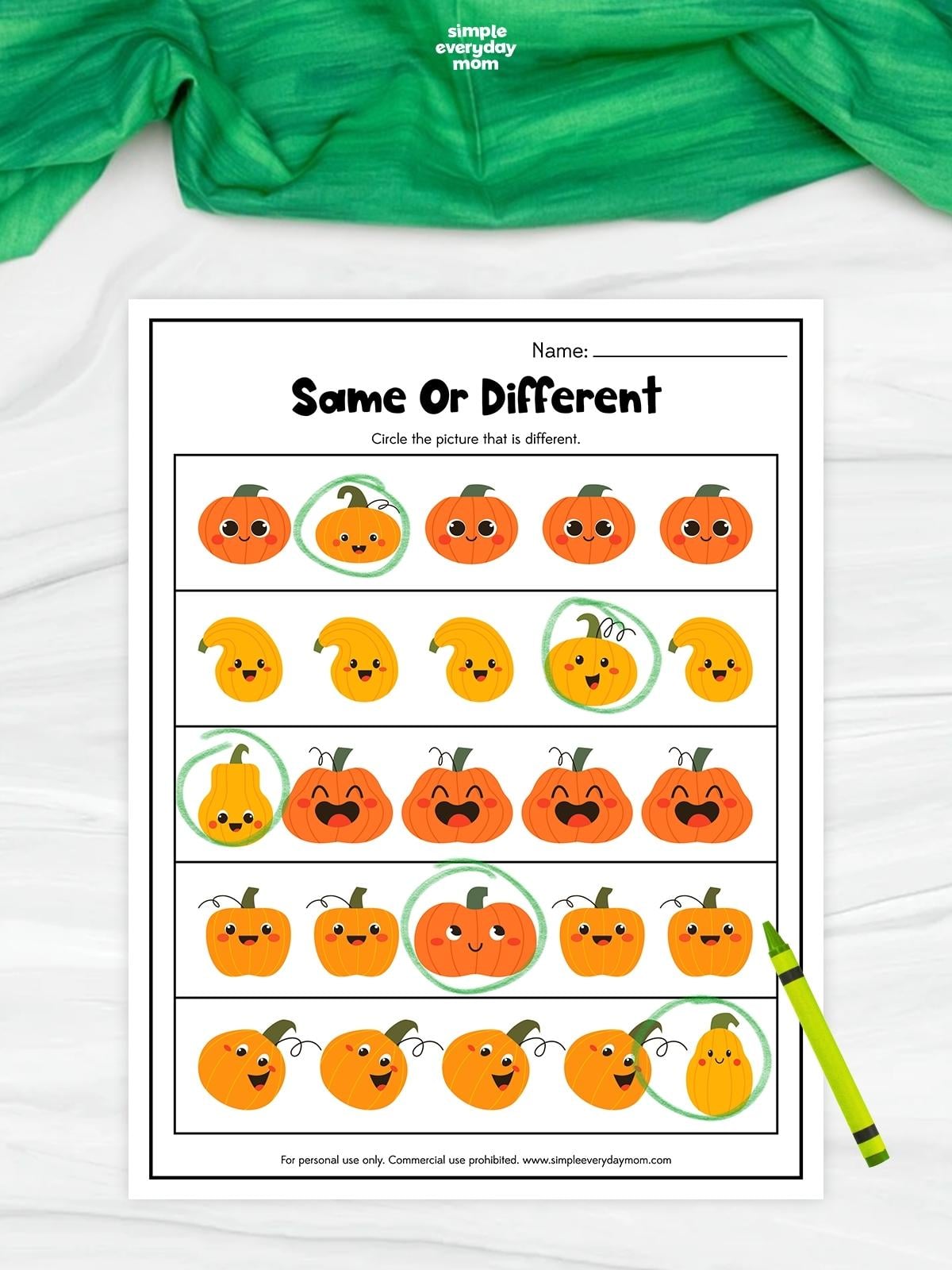 A completed same or different pumpkin worksheet features rows of cheerful pumpkins and gourds. The picture that is different in each row is circled in green to practice visual discrimination. A green crayon rests beside the printable on a white surface with green fabric above.