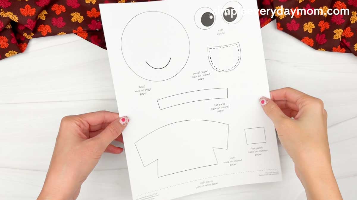 Hands holding the printable Fork Painted Scarecrow Craft template with the head, shirt, hat band, eyes, pocket, and hat patch pieces.