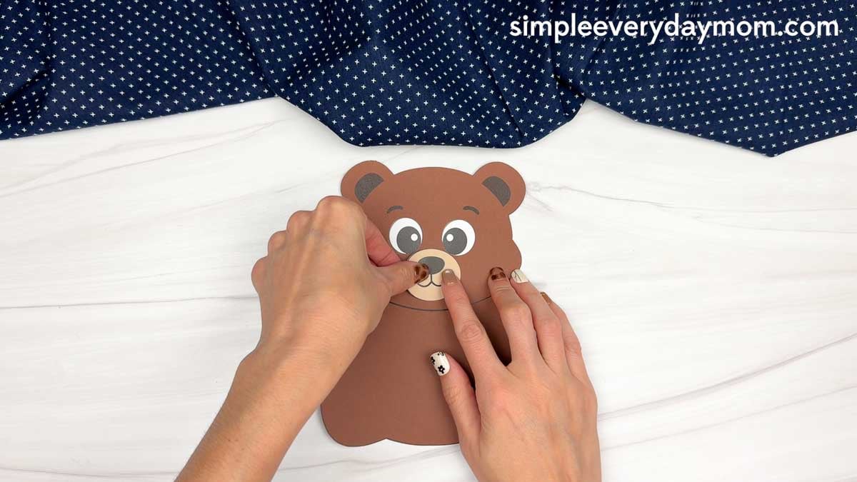 hands attaching the muzzle and nose piece to a brown paper bear craft.