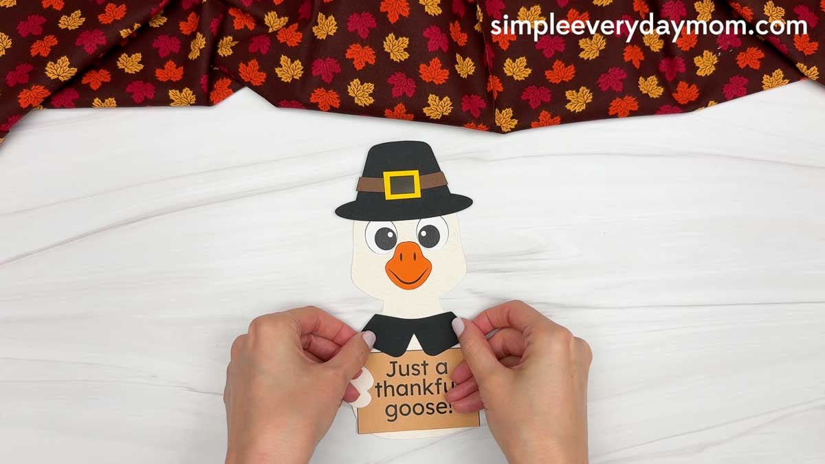 Hands gluing a black collar below the goose's head and above the thankful sign for a Thanksgiving paper craft.