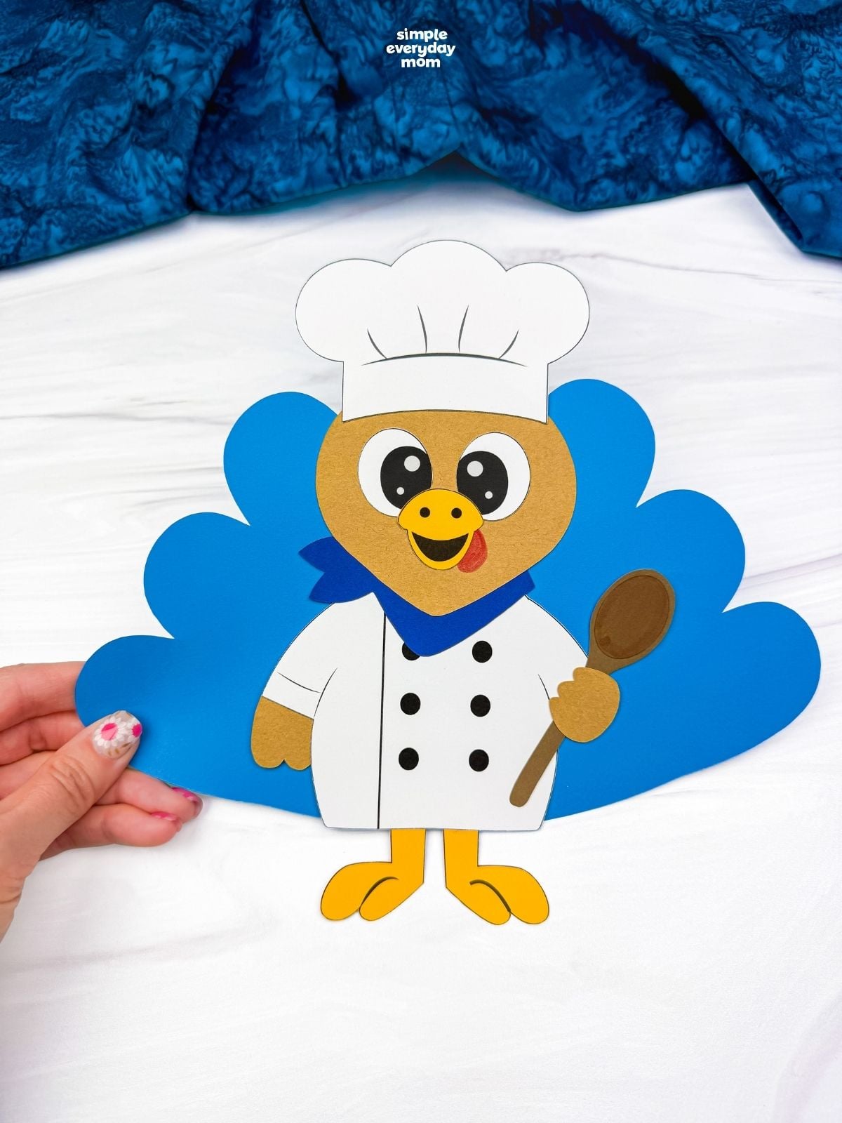 hands holding a completed chef turkey disguise craft with blue tail feathers and a brown spoon.