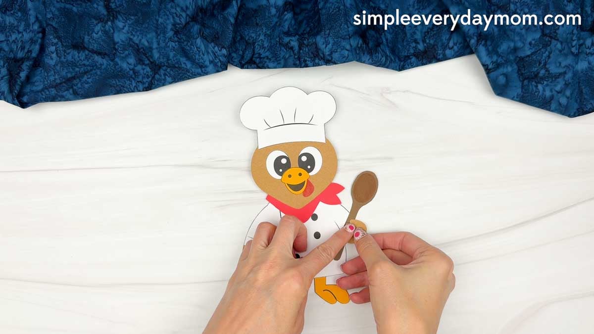 hands attaching a brown spoon to the chef turkey disguise craft.