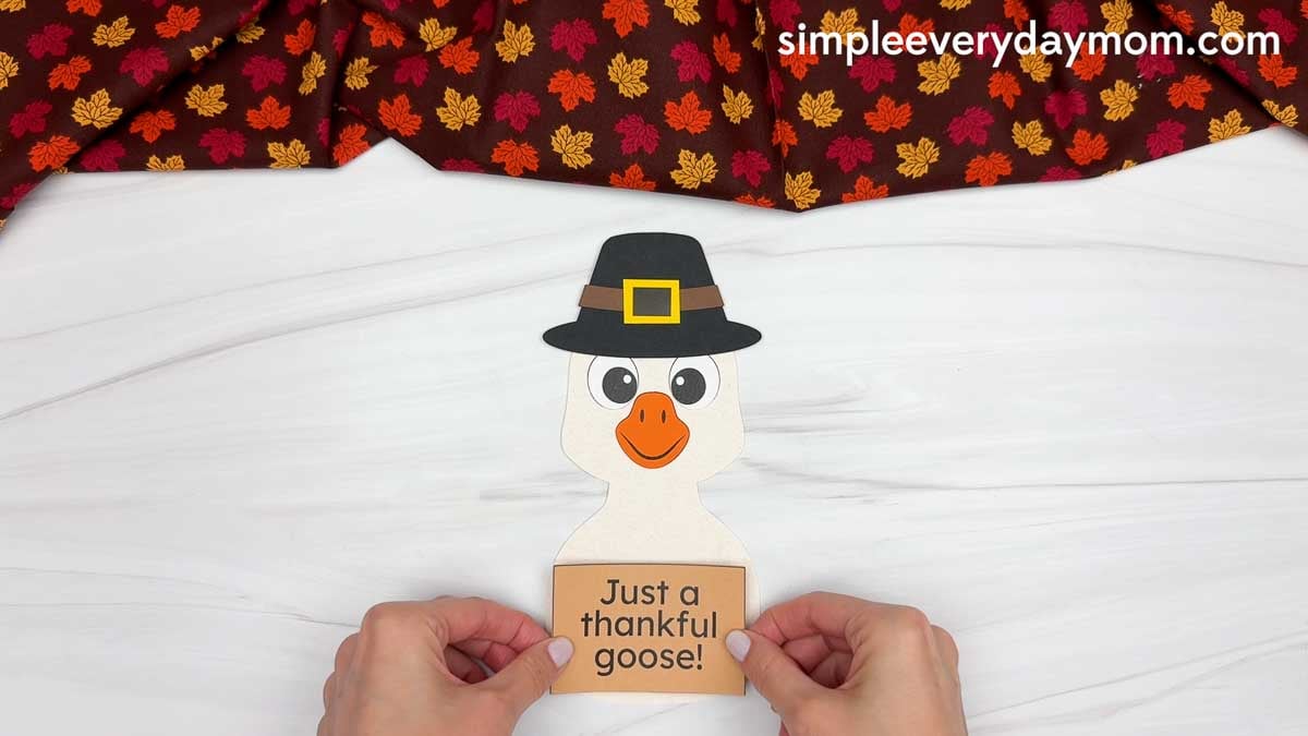 Hands placing a "Just a thankful goose!" sign on the goose's body for a Thanksgiving paper craft.