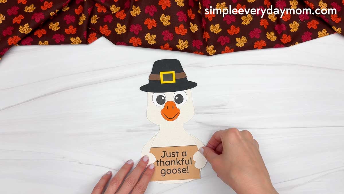 Hands gluing the goose's wings on each side of the thankful sign for a Thanksgiving paper craft.