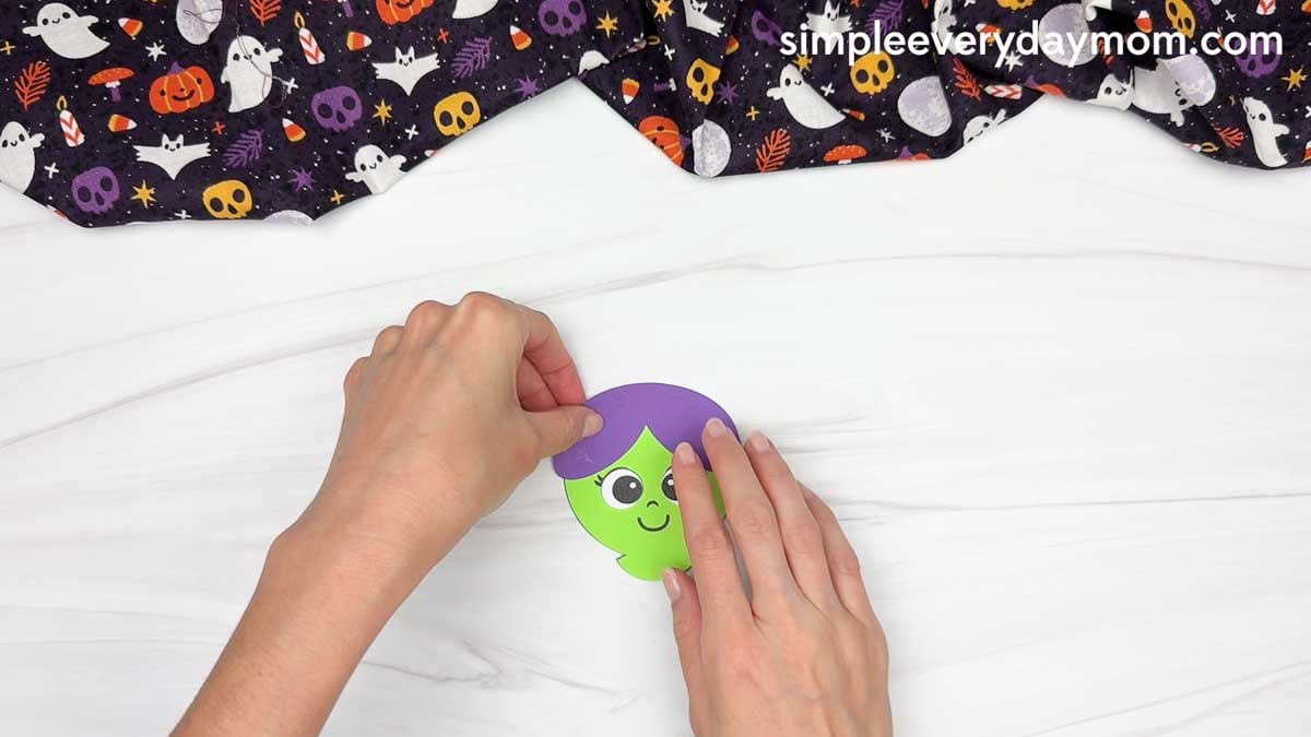 Hands gluing the purple hair onto the top of the witch’s face.