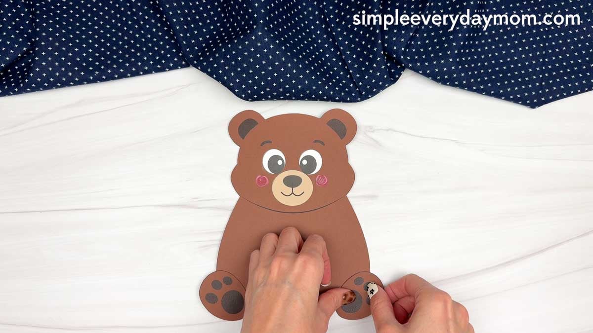 hands attaching a paw piece to the bottom of a brown paper bear craft.