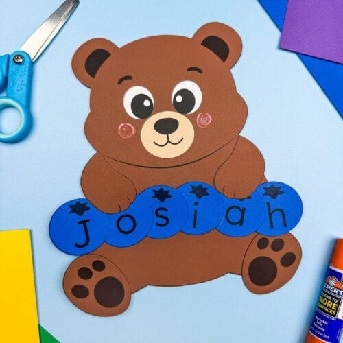 A completed Craft featuring a brown bear holding blue letter pieces spelling Josiah, surrounded by colorful craft supplies.