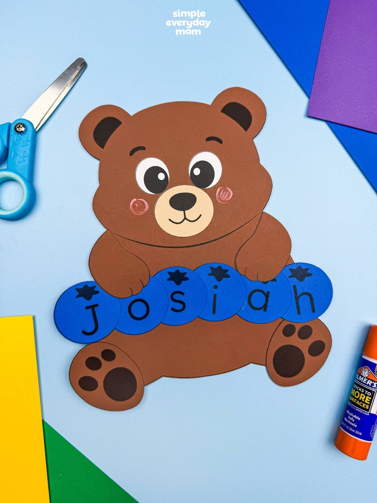 A completed Craft featuring a brown bear holding blue letter pieces spelling Josiah, surrounded by colorful craft supplies.