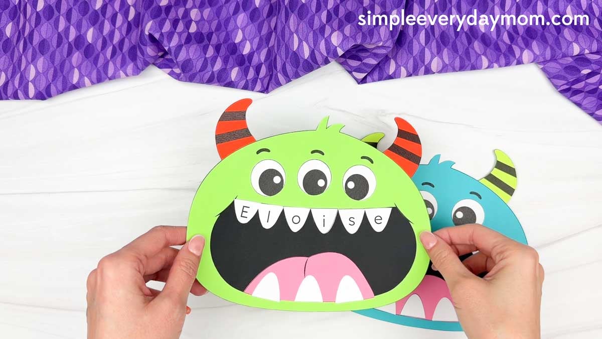Hands holding a completed monster name craft with letters written across the monster’s top teeth.