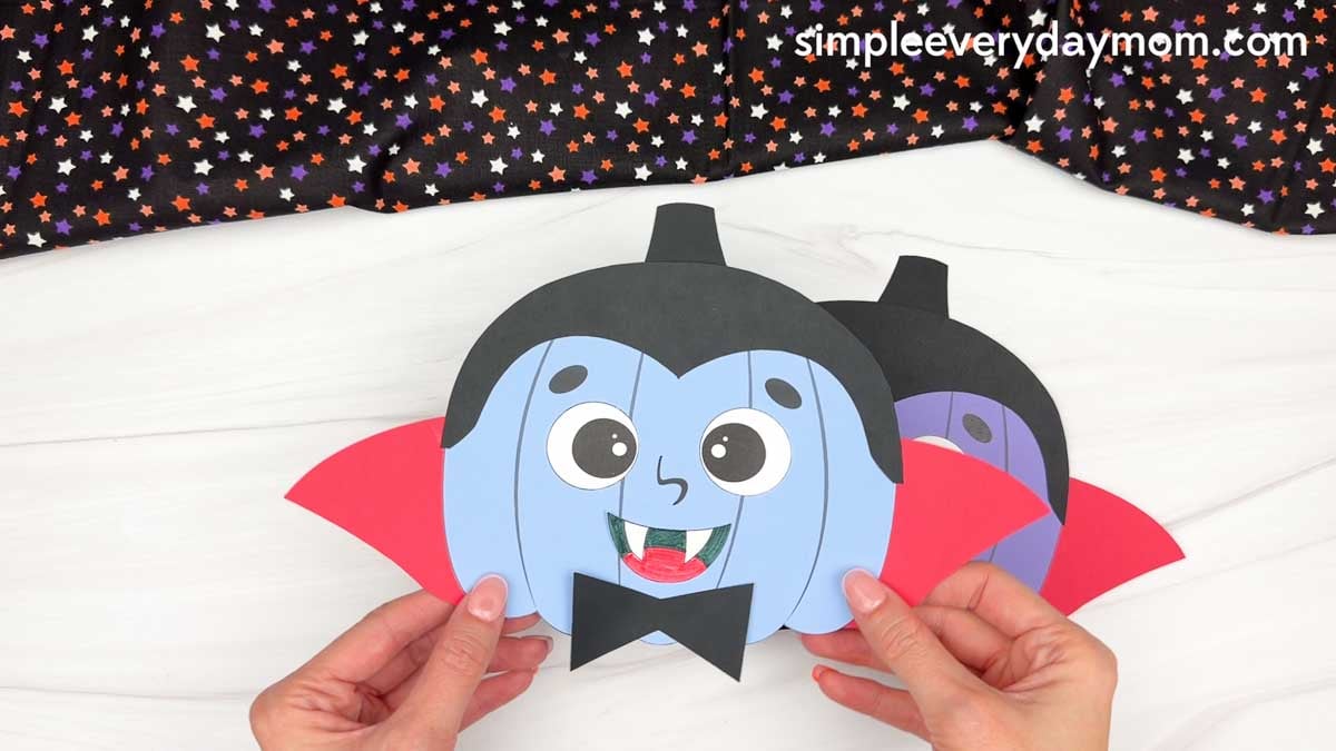 Hands holding a completed vampire pumpkin craft with a cape, fangs, bow tie, and black hair.