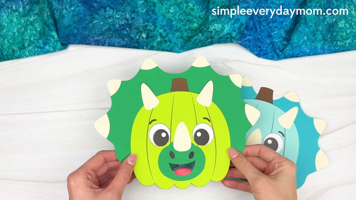 Hands holding a completed green dinosaur pumpkin craft with horns, a smiling face, and a spiked frill.