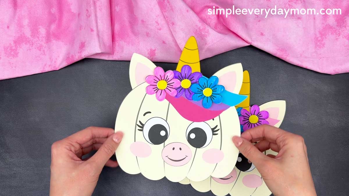 Hands holding a completed unicorn pumpkin craft decorated with a horn, ears, colorful flowers, and a flower crown.