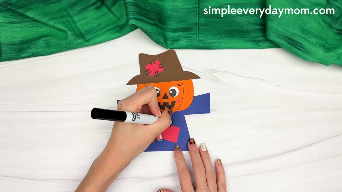 Hands drawing details on the scarecrow body below the pumpkin head.