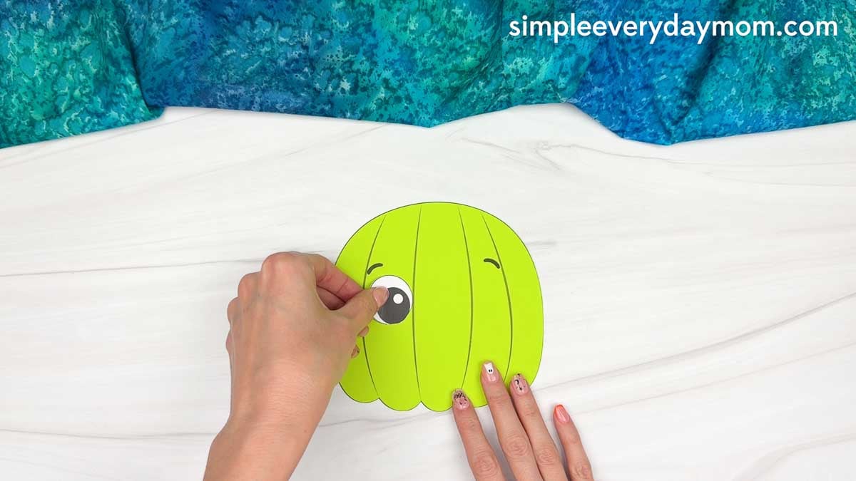Hands gluing an eye onto the upper left side of the dinosaur pumpkin craft.