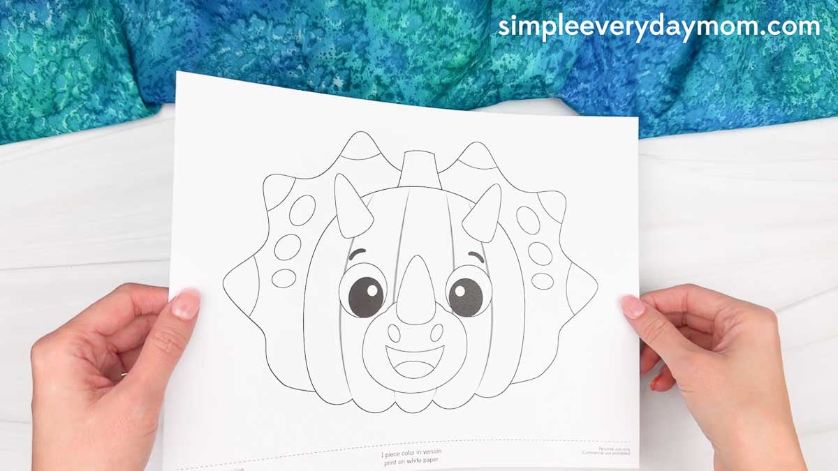 Hands holding a black and white dinosaur pumpkin craft template ready for kids to color.