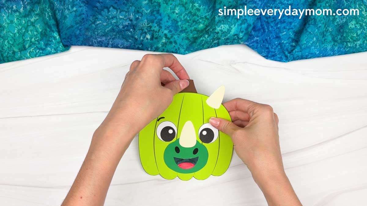 Hands gluing the stem onto the top of the dinosaur pumpkin craft.