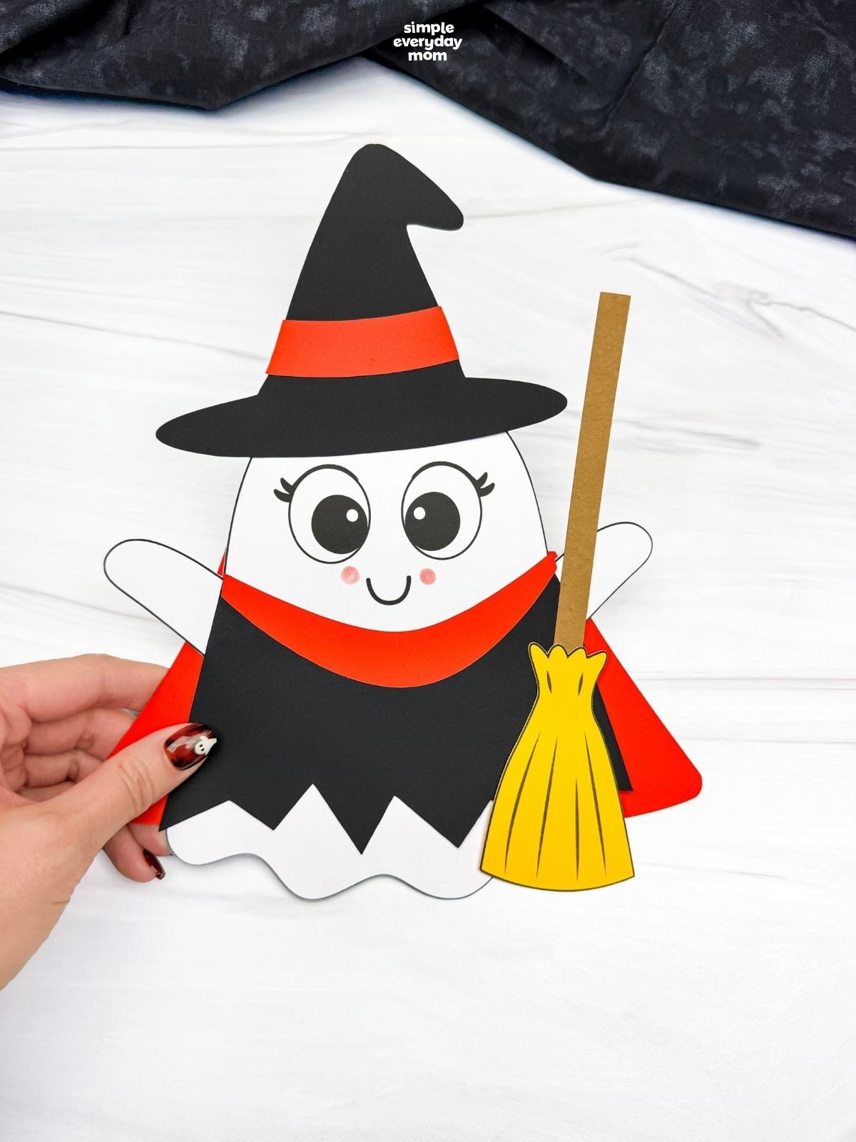 Hands holding a completed Witch Ghost Disguise craft with a witch hat, dress, and broom.