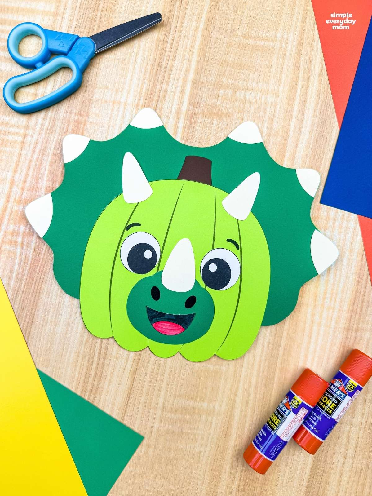 A completed craft showing a green dinosaur pumpkin disguise with horns and a spiked frill beside craft supplies.