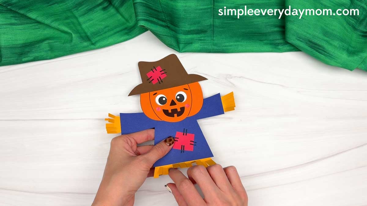Hands holding a completed pumpkin head scarecrow with a patched shirt and straw details.