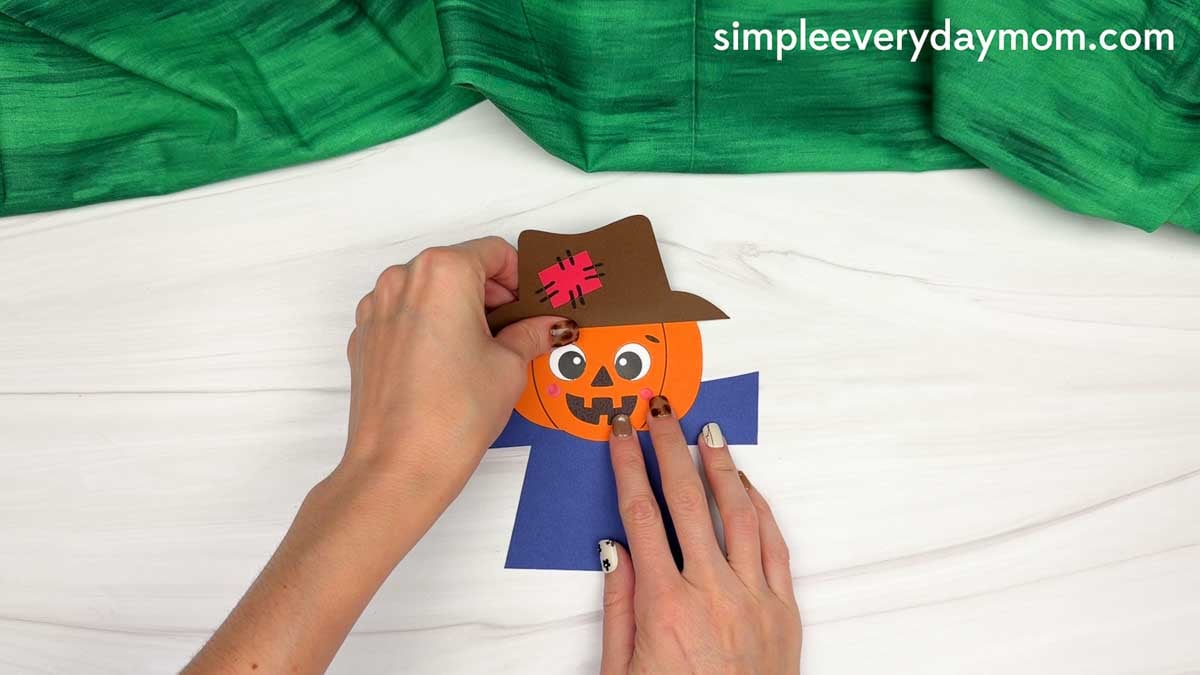 Hands attaching the pumpkin head to the top of the scarecrow body.