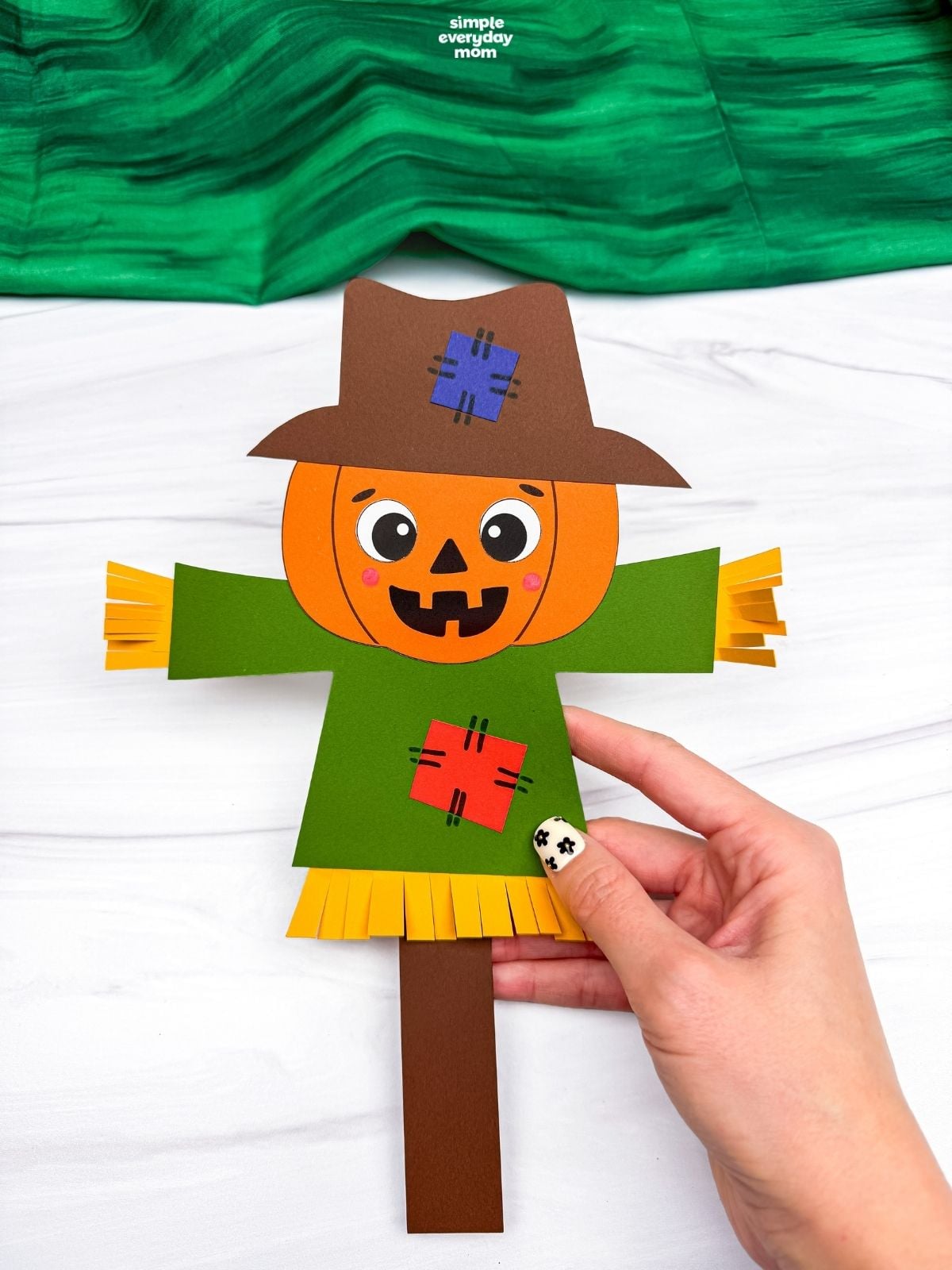 Hands holding a completed pumpkin head scarecrow with a patched hat, shirt, and straw details.
