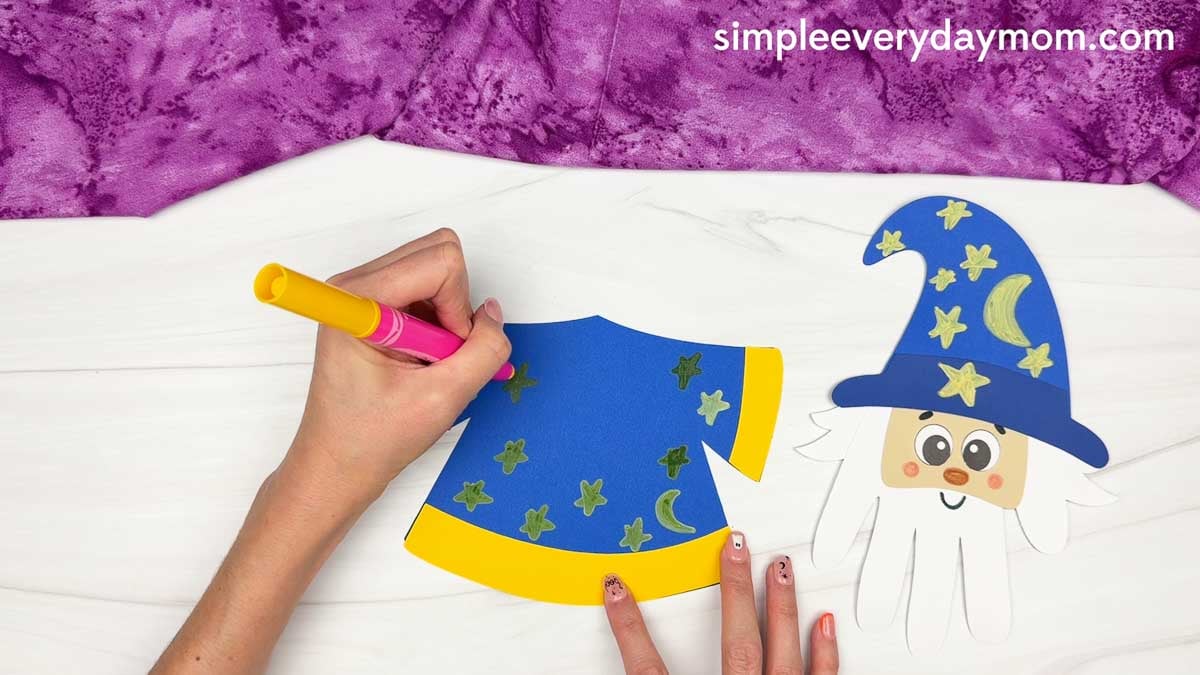 Hands decorating the wizard robe with stars and a moon for the Wizard Handprint Craft.