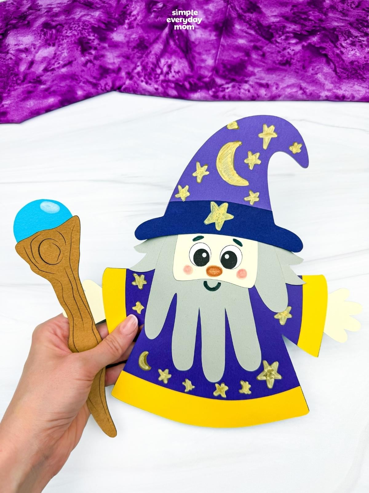 Hands holding a completed Wizard Handprint Craft with a decorated hat, handprint beard, robe, and wizard staff.