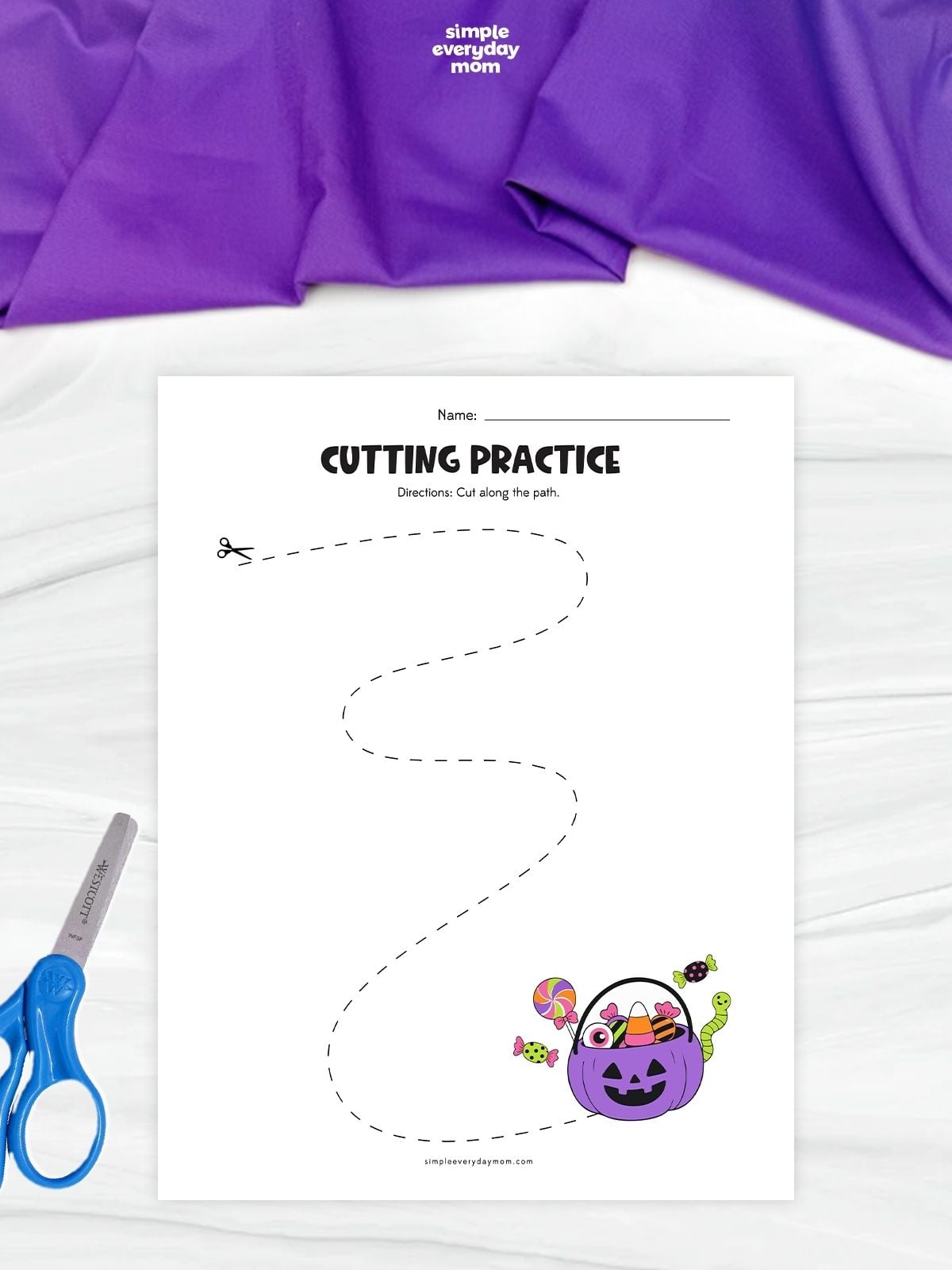 Halloween-themed cutting practice worksheet with a long dashed, winding path leading to a purple jack-o’-lantern bucket filled with colorful candy.