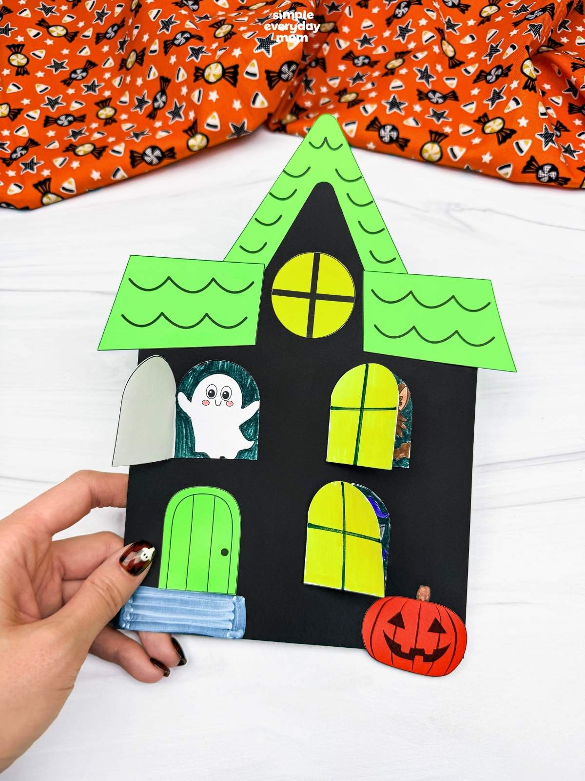 Hands holding a completed Peekaboo Haunted House Craft with opening windows revealing Halloween characters, a door, and a pumpkin.