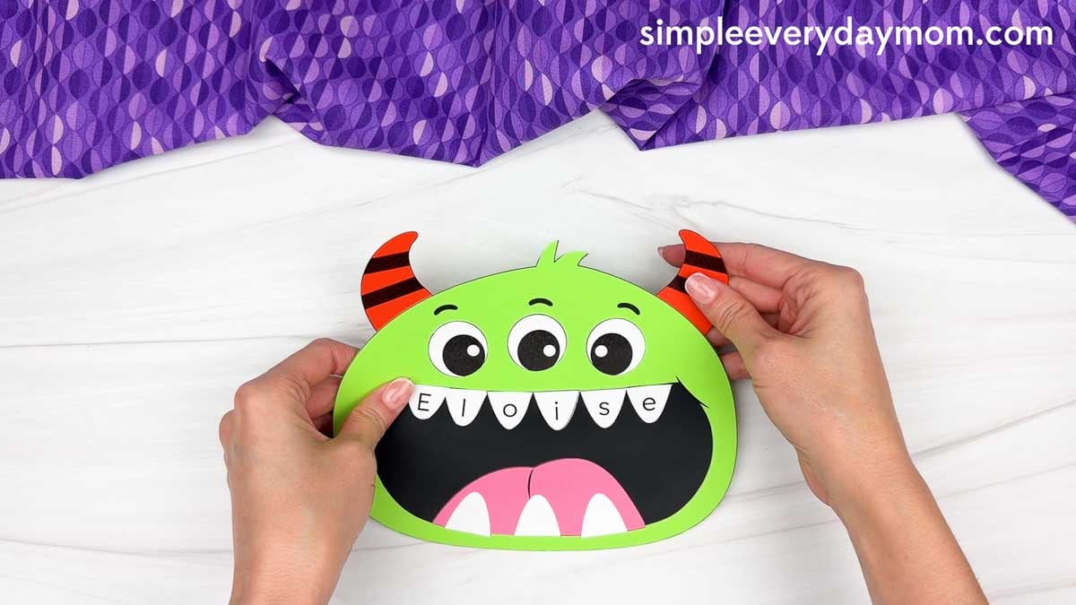 Hands gluing a striped horn to the top right side of the monster’s head.