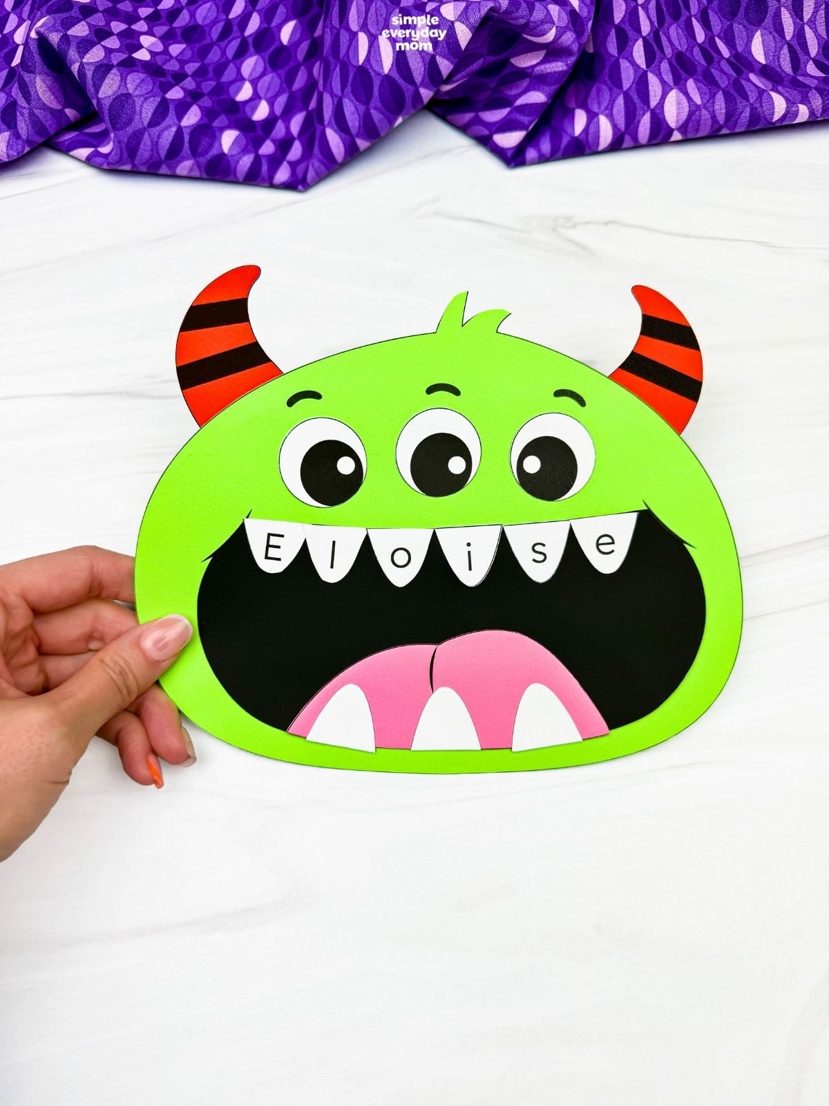Hands holding a completed green monster name craft with three eyes, striped horns, and the name Eloise written across its teeth.