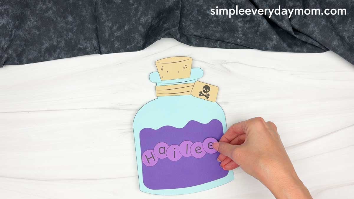 Hands adding letter bubbles that spell a name across the Halloween potion bottle craft.