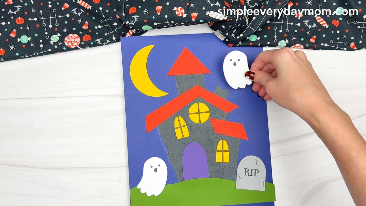 Hands placing a ghost above the torn paper haunted house scene.