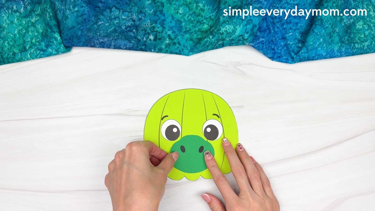 Hands gluing the snout onto the center of a green dinosaur pumpkin craft.