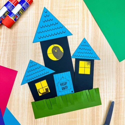 A completed craft showing a haunted house made from paper shapes with triangular roofs, square windows, a round window, door, and grass.