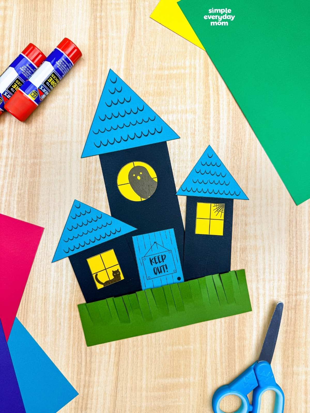 A completed craft showing a haunted house made from paper shapes with triangular roofs, square windows, a round window, door, and grass.