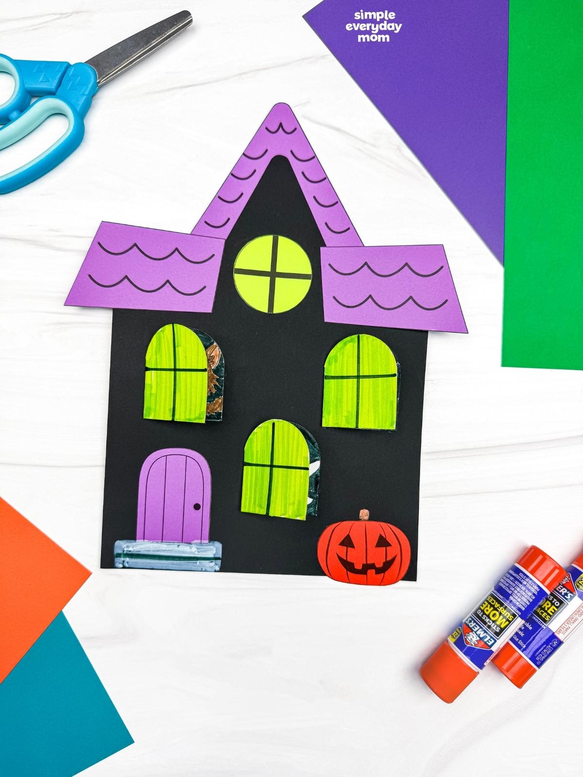 A completed craft featuring a Peekaboo Haunted House with opening windows, a colorful roof, door, and jack-o'-lantern.