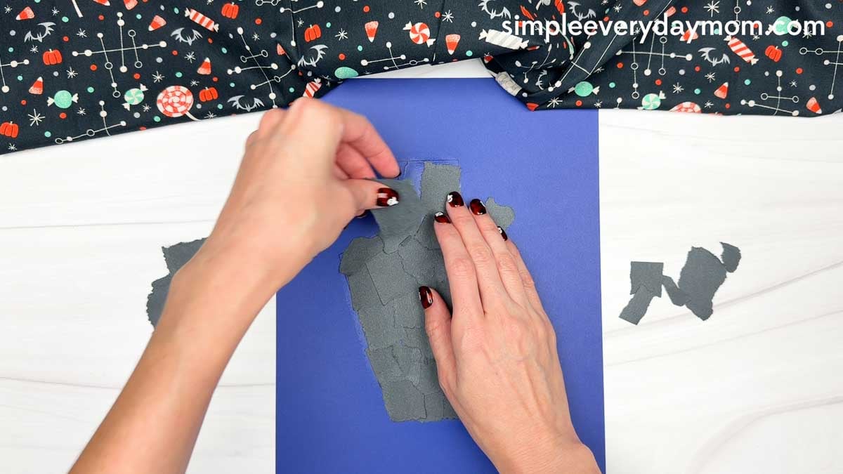 Hands tearing gray paper into small pieces to create the haunted house on a dark blue background.