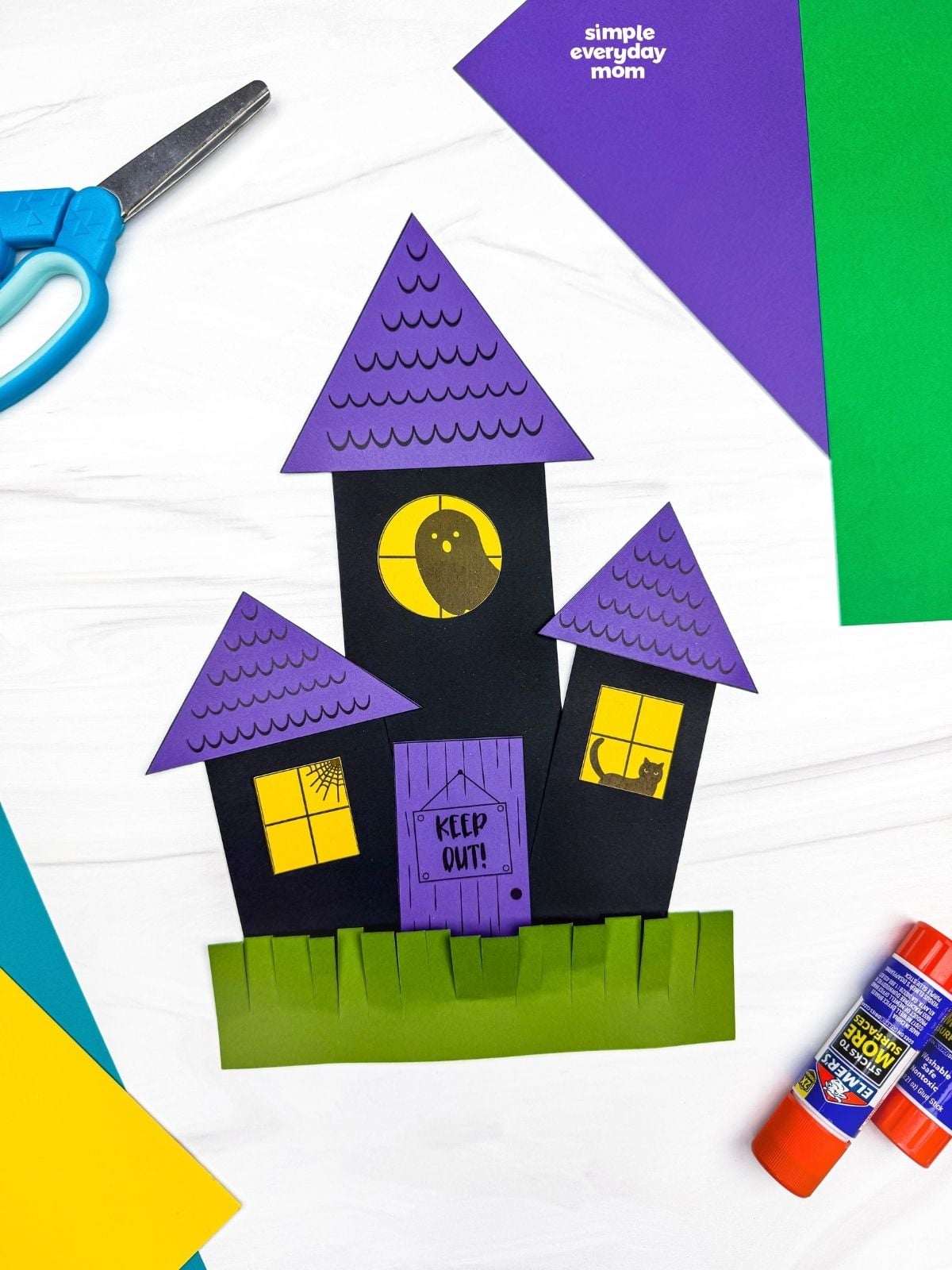 A completed craft showing a haunted house made from paper shapes with triangular roofs, windows, a door, and fringed grass.
